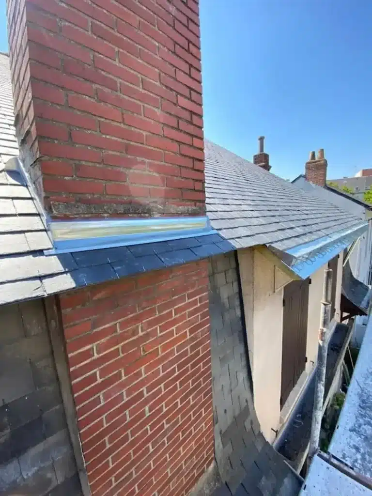 Étanchéité de cheminée avec solin neuf sur toit ardoise Travaux de zinguerie sur une cheminée en brique rouge avec solin métallique neuf et toiture en ardoises grises.